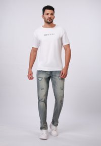 Weißes Baumwoll-T-Shirt mit japanischem Text, kombiniert mit hellblauen verwaschenen Jeans und weißen Sneakers, vor einem neutralen Hintergrund stehend.