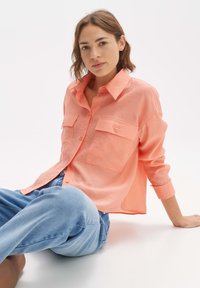 Licht koraalkleurig overhemdd shirt met twee borstzakken, lange mouwen en een relaxte pasvorm. Geselecteerd met blauwe jeans tegen een effen achtergrond.