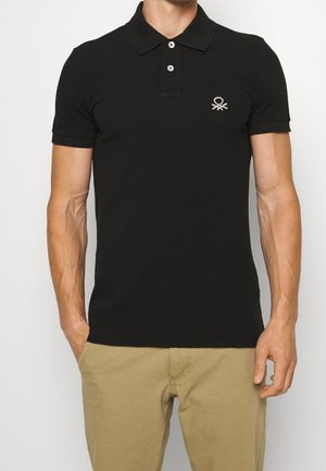 Hombre vistiendo una camiseta tipo polo negra ajustada con dos botones y un pequeño logo bordado blanco en el pecho, combinado con pantalones beige.