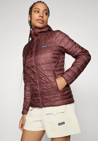 Femme portant une veste matelassée bordeaux avec une capuche et un short beige, regardant vers sa gauche avec une main dans la poche de la veste.