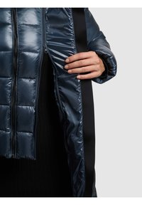 Hånd trækker en mørkeblå quiltet jakke op, der viser sort inderbeklædning og lynlåsdetalje på lys grå baggrund.