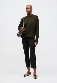 Suéter verde escuro com decote canelado, combinado com calças pretas ajustadas e cropped. Sapatos slip-on de padrão leopardado e uma pequena bolsa preta cruzada.