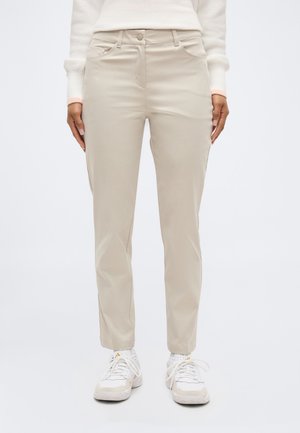 Άτομο που φοράει μπεζ παντελόνι ίσιας γραμμής, λευκά αθλητικά παπούτσια και λευκό πουλόβερ, στέκεται με τα χέρια χαλαρά δίπλα στο σώμα.