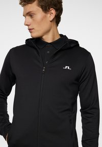 Svart zip-up hoodie med hög krage, integrerad huva och en vit logotyp på bröstet. Slät tyg med en åtsittande design.