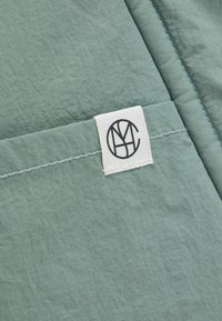 Κοντινή λήψη ενός ανοιχτοπράσινου υφάσματος με ραμμένες ενώσεις και μια λευκή ορθογώνια ετικέτα που φέρει μαύρο κυκλικό λογότυπο μονογράμματος.