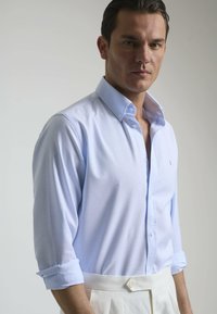 Camisa de botones azul claro con un emblema en el pecho, puños enrollados y textura suave, combinada con pantalones crema.