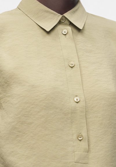 Chemise beige boutonnée avec un col classique, portée par une personne à la peau foncée, montrant la zone du milieu de la poitrine et du cou.