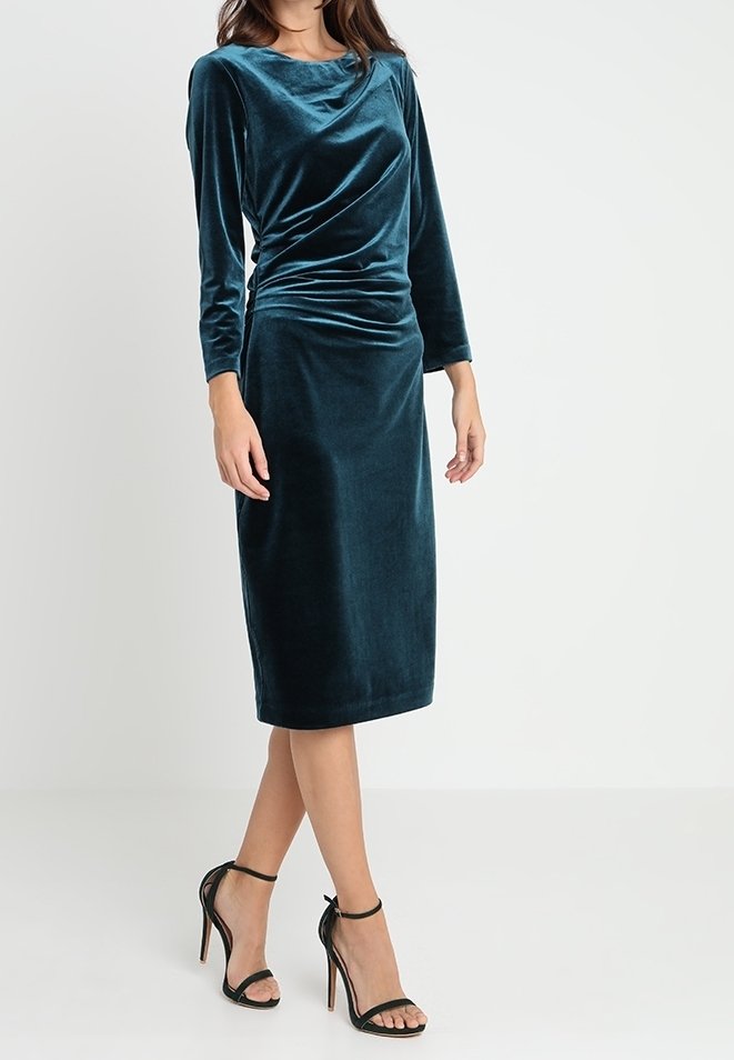 Petrolblå sammetklänning med långa ärmar och draperad framsida, knälång nederkant, kombinerad med svarta sandaletter med hög klack och remmar.