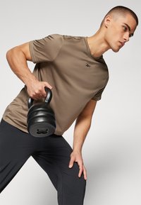 T-shirt de sport marron et pantalon de sport noir, tenant un kettlebell noir. Design lisse et ajusté ; branding minimal élégant visible.