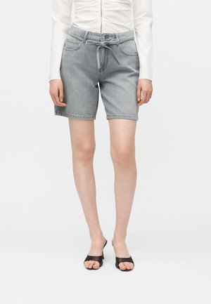 Personne portant un short en jean gris mi-long avec une taille à cordon, un haut blanc à manches longues froncé et des sandales à talons ouvertes noires.