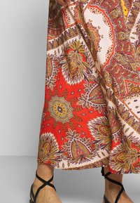 Jupe longue à base rouge vif ornée de motifs en paisley et floraux complexes en jaune, violet et crème, fabriquée en tissu léger.