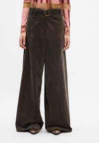 Pantalons en velours côtelé à jambes larges de couleur marron foncé, taille haute avec une boucle de ceinture circulaire dorée. Texture douce et coupe structurée.