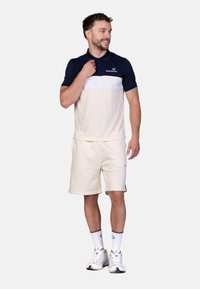 Poloshirt met korte mouwen in marineblauw en crème, met een horizontale witte streep, gecombineerd met lichtcrème shorts en witte sneakers.