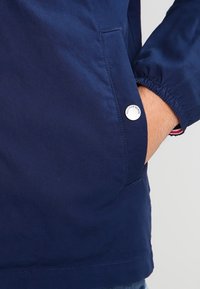 Marinblå jacka med en slät, matt yta. Har en sidoficka med en metallknapp och elastiska ärmslut. Enkelt design, inga mönster.