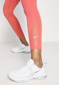 Korale legginsy fitnessowe z siateczkowymi akcentami i białym logo Nike, w połączeniu z białymi butami sportowymi z srebrnymi detalami i oddychającą siateczką.