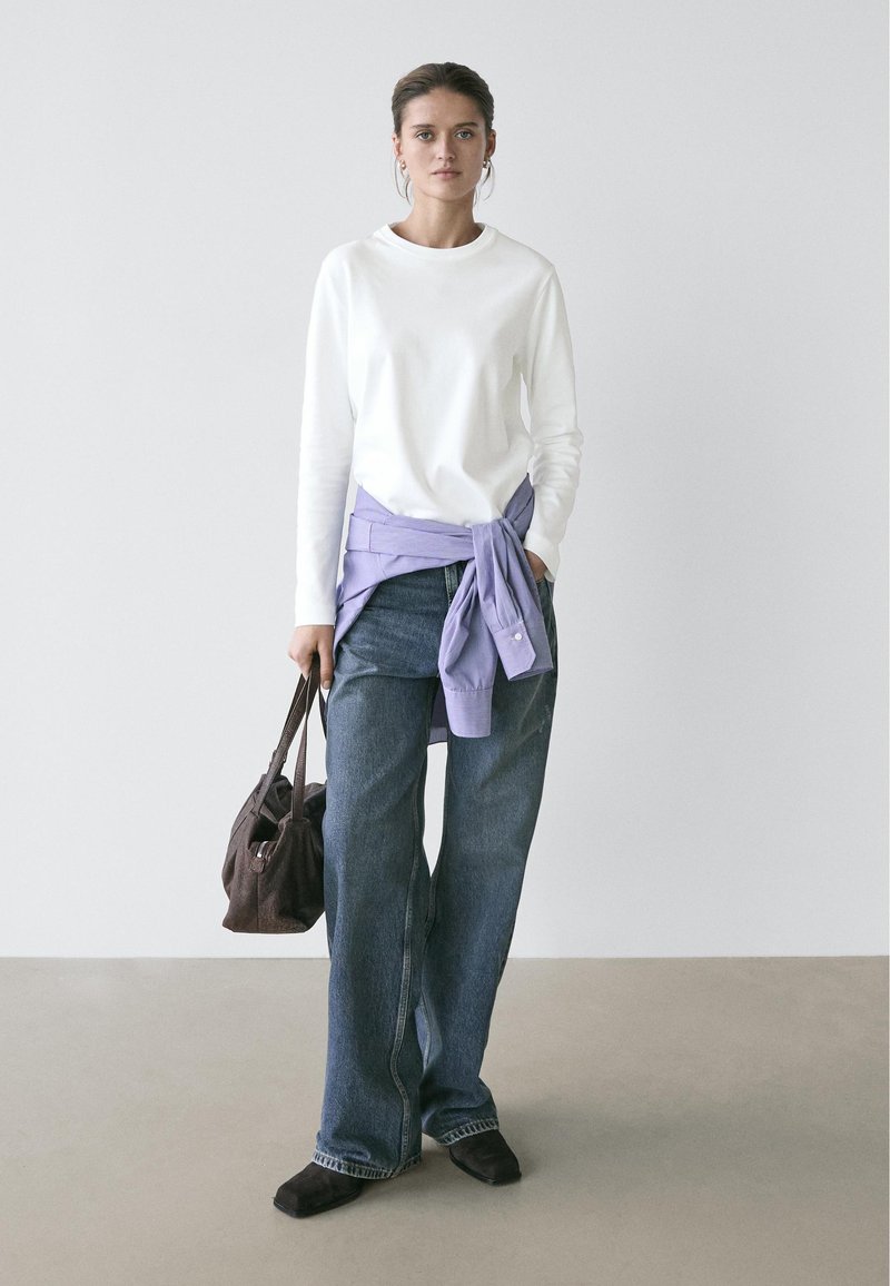 Femme debout portant un haut blanc à manches longues, un jean bleu, une chemise violette nouée à la taille, des chaussures noires, tenant un sac à main marron foncé dans un espace neutre.