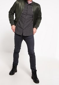 Oliivinvärinen bomber-takki ruudullisen musta-harmaan kauluspaidan päällä, yhdistetty tummiin kapeisiin farkkuihin ja mustiin nyörisaappaisiin. Sileitä, teksturoituja materiaaleja.