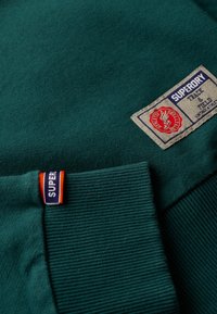 Felpa verde con tessuto testurizzato, polsini a costine e un'etichetta tessuta con logo e testo in colori a contrasto.