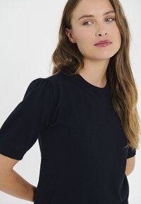 Jūras zilas krāsas īsām piedurknēm ražots topiņš no mīksta materiāla, ar apaļu dekoltē un pūkainām piedurknēm, lai pievienotu tekstūras detaļu.