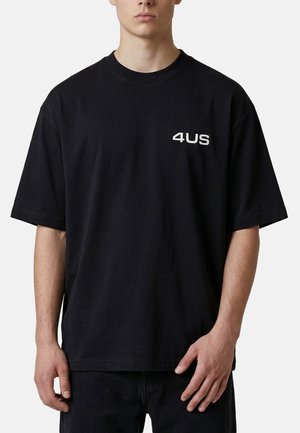 Giovane uomo che indossa una t-shirt nera oversize con scritta bianca "4US" sul petto sinistro, in piedi davanti a uno sfondo semplice.