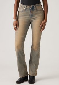 Άτομο που φοράει ξεθωριασμένα μπεζ τζιν με ευθείς γραμμές και μαύρα μποτάκια με τακούνι στον αστράγαλο, με ασημένια δαχτυλίδια και βραχιόλια στα χέρια.