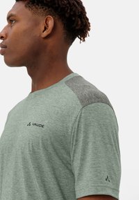 Camicetta verde chiaro a maniche corte con dettagli grigi sulle spalle; presenta un logo sul petto, una texture morbida e una vestibilità casual.