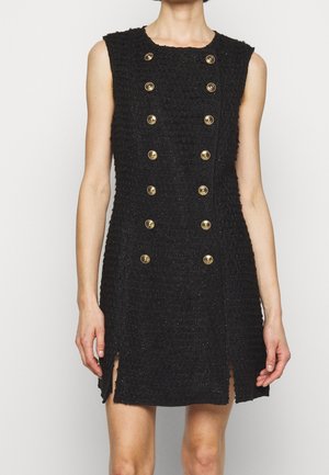 Robe noire sans manches à texture, avec double rangée de boutons décoratifs dorés et petites fentes à l'avant, portée par une personne debout.