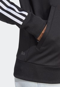 Hand i en svart jacka med vita ränder på ärmen glider ner i framfickan, tillsammans med ljusblå jeans.