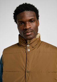 Brun vadderad väst med hög krage, prydd med metallknappar med "Lee"-logotyp, kombinerad med en denimskjorta under.