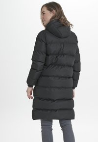 Abrigo acolchado negro con capucha, con secciones acolchadas horizontales y puños elásticos. Llevado sobre unos jeans grises, visto desde la parte de atrás.