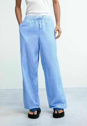 Persoon draagt lichtblauwe en witte verticale gestreepte wijde broek met koordsluiting in de taille, wit hemdje en zwarte platform sandalen.