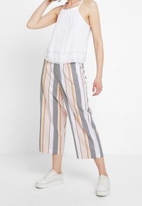 Haut blanc sans manches avec un détail en dentelle à l'ourlet, associé à un pantalon crop à rayures multicolores dans des tons pastel et des baskets blanches.