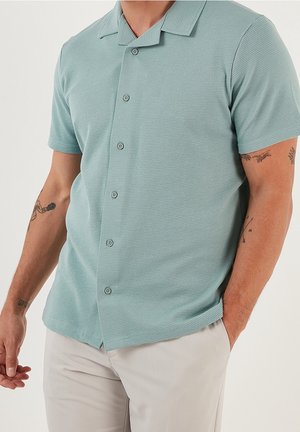 Mann trägt hellblaues, kurzärmeliges Hemd mit Knopfleiste und Kragen, beige Hose und sichtbare Tattoos auf beiden Unterarmen.