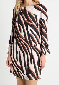 Robe à manches longues avec un motif abstrait à rayures crème, noir et rouille. Présente une coupe droite et des poignets boutonnés. Tissu à texture lisse.