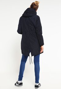 Černá prošívaná parka s kapucí, staženým pasem a elastickými šňůrkami. Obsahuje dlouhé rukávy a rozparek vzadu pro snadnější pohyb.