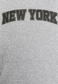 Sivá mikina s okrúhlym, čiernym nápisom "NEW YORK" cez hruď. Materiál vyzerá mäkký s jemnou textúrou. Obsahuje predné vrecko.