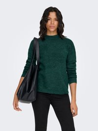 Femme aux cheveux ondulés et foncés, portant un pull vert foncé et un pantalon noir, avec un grand sac à bandoulière en cuir noir.