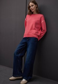 Femme appuyée contre un mur sombre, portant un pull corail à manches longues, un jean large bleu foncé et des baskets beiges, regardant légèrement sur le côté.