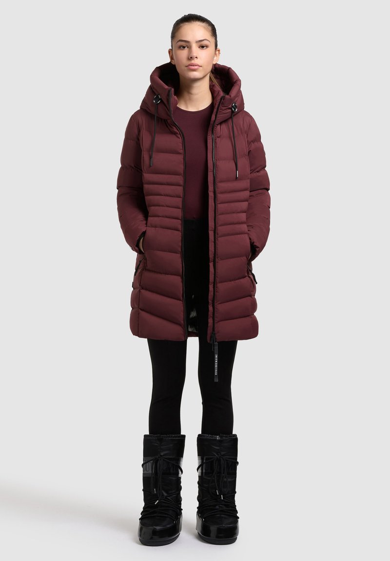 Maroon puffer jacket with a hood, featuring horizontal quilted design, front zipper, and side pockets. Worn over black leggings and boots.
