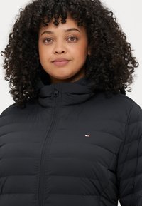 Schwarze gepolsterte Jacke mit hohem Kragen und Reißverschluss. Verfügt über ein rotes, weißes und blaues Logo auf der Brust. Textil mit strukturiertem, gestepptem Design.