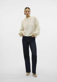 Pull en maille crémeux avec un col large et des manches volumineuses, associé à un jean droit bleu foncé et des chaussures à talons beiges.