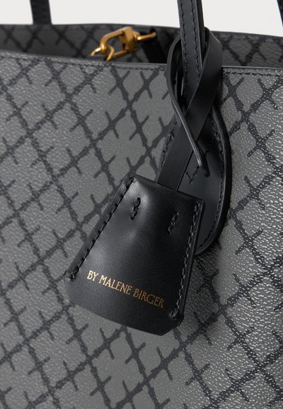 Gros plan d’un sac à main gris texturé avec motif croix noir, étiquette en cuir noir et texte doré indiquant "BY MALENE BIRGER."