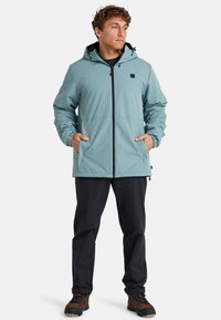 Chaqueta azul clara con capucha, puños elásticos, cierre frontal con cremallera y bolsillos laterales; combinada con pantalones negros y botas de senderismo marrones.