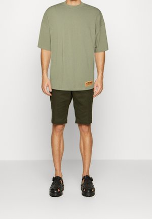 Olivegrünes Baumwoll-T-Shirt mit kurzen Ärmeln und einem rechteckigen Patch-Label, kombiniert mit dunkelgrünen Shorts und schwarzen Sandalen.