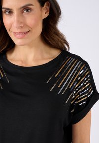 Zwarte t-shirt met korte mouwen en een ronde halslijn, versierd met metallic pailletten in goud, blauw en zilver, gerangschikt in diagonale strepen op de schouders.