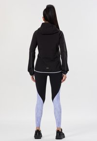 Giacca nera con cappuccio e orlo elastico, abbinata a leggings neri e blu chiaro con motivi. L'outfit presenta un design elegante e dettagli funzionali.