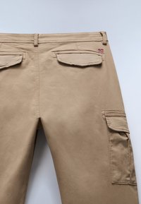 Beige cargo bukser med en glat tekstur har to baglommer og sidelommer til cargo, forstærkede sømme og et lille emblem på livet.
