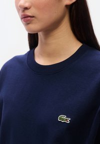 Μπλε ναυτικό βαμβακερό φούτερ με στρογγυλή λαιμόκοψη, ριμπ μανσέτες και μικρό πράσινο κεντημένο λογότυπο κροκόδειλου στο στήθος.