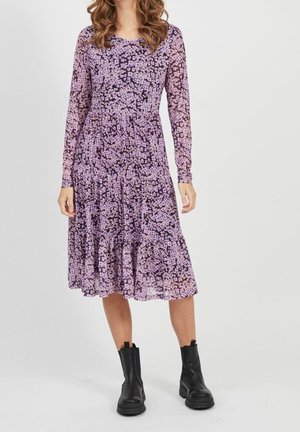 Femme portant une robe midi violet à fleurs à manches longues, associée à des bottines noires, debout devant un fond clair uni.