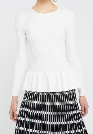 Kobieta ubrana w biały prążkowany top z długim rękawem i dołem w stylu peplum oraz plisowaną spódnicę w czarno-białe pionowe paski.